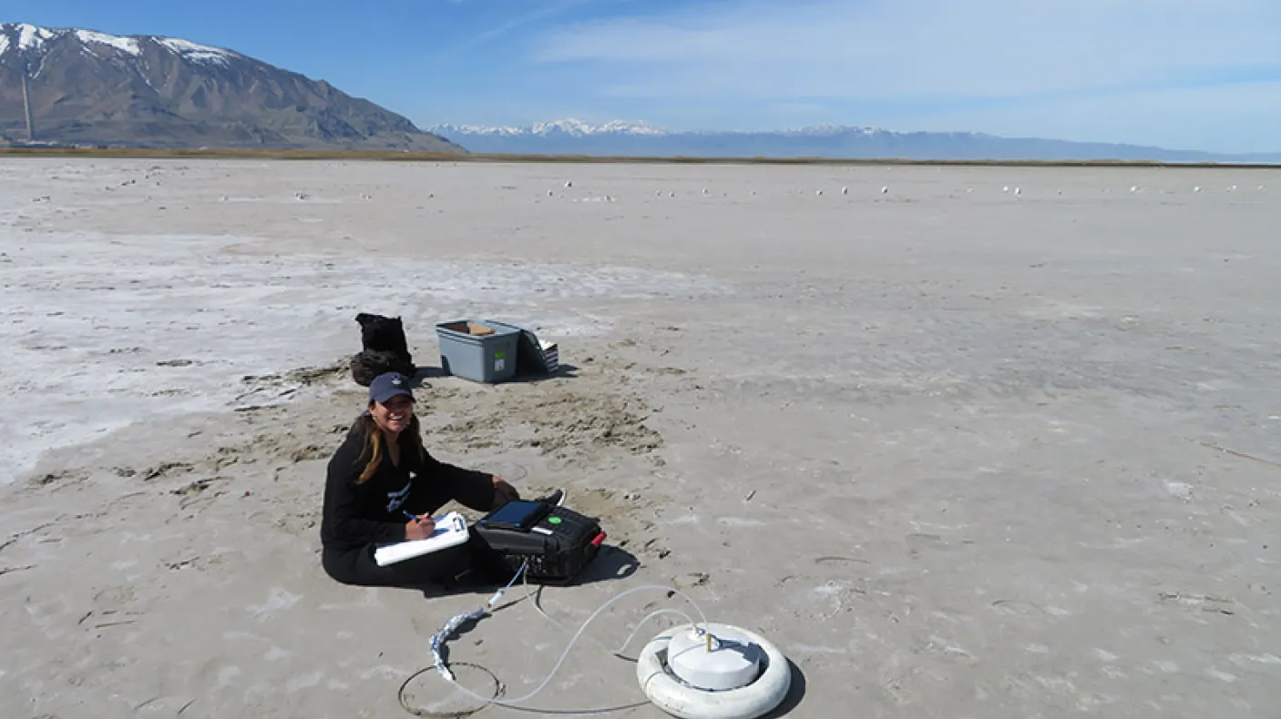 Researcher sitting on a dry lakebed next to scientific equipment including a circular white sensor device.