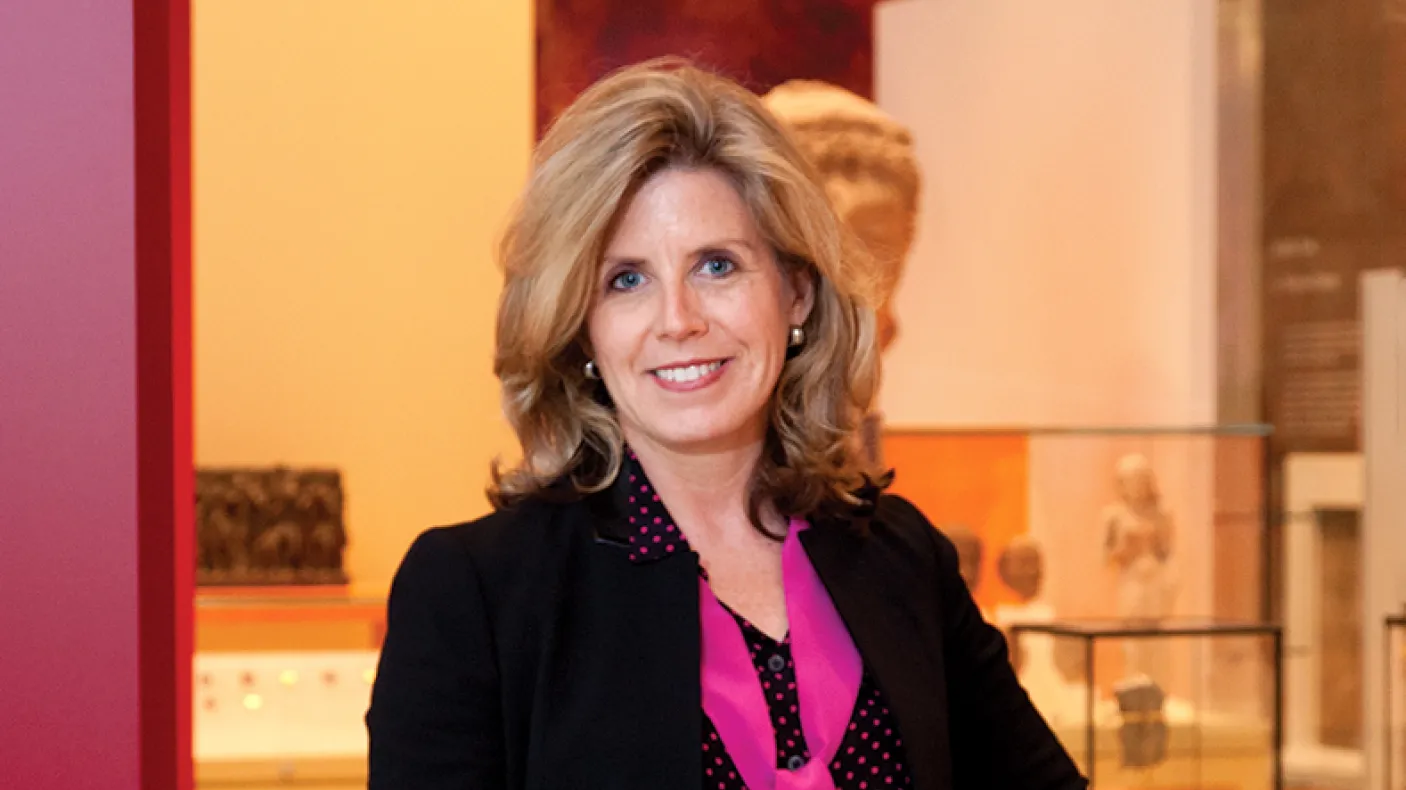 Woman with shoulder-length blonde hair wearing a black blazer over a pink blouse, standing indoors with blurred background.
