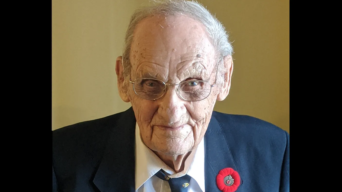 Elderly man wearing a dark blazer, striped shirt, and tie with a red poppy pin on his lapel.