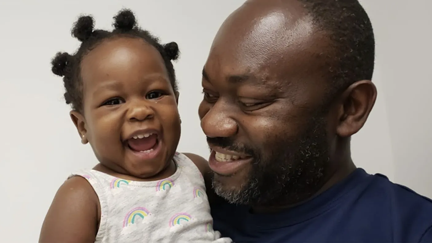 A smiling man in a blue shirt holding a laughing toddler wearing a white tank top with rainbow patterns.