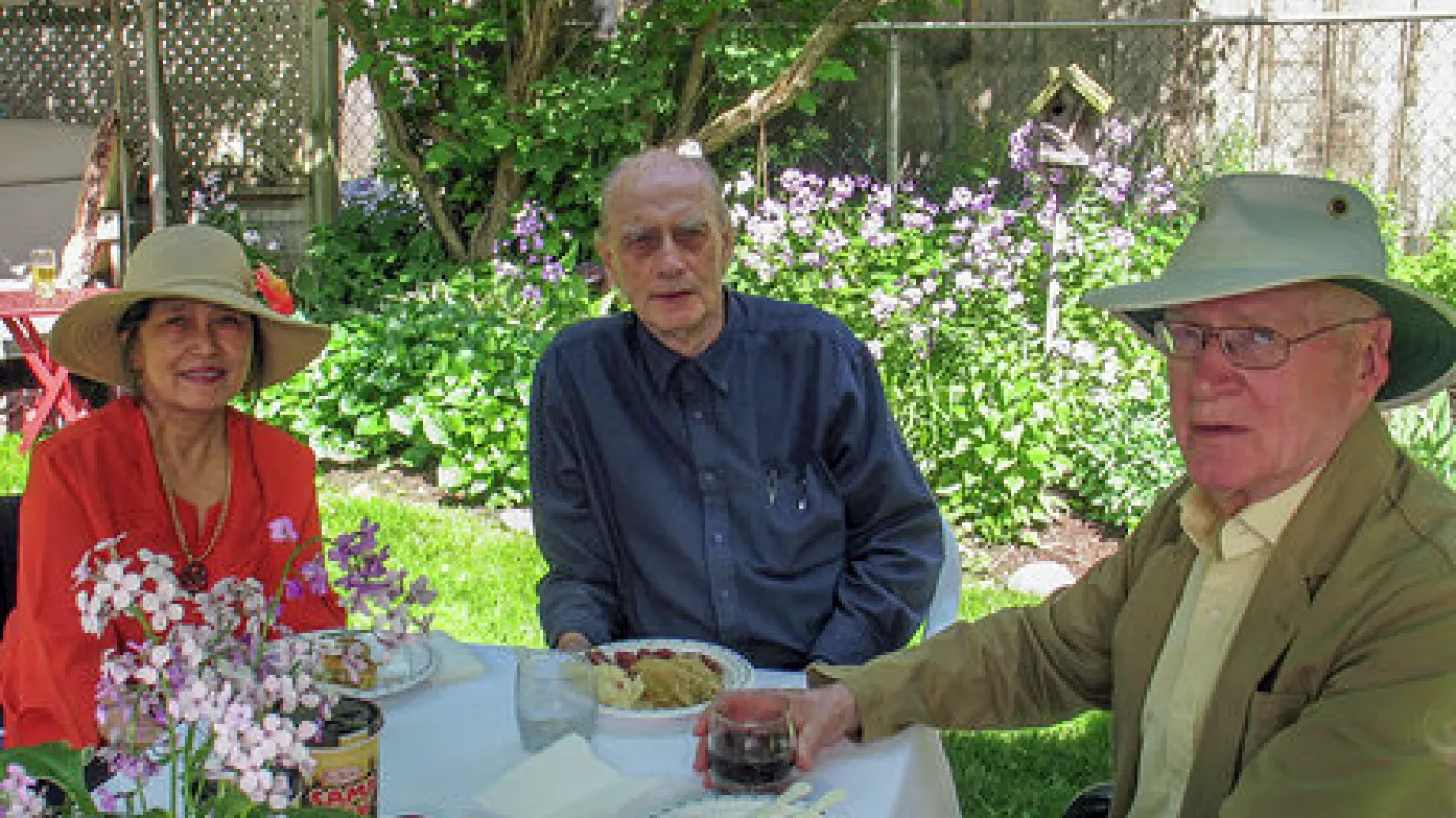 Three older adults sitting outdoors at a table with flowers and greenery in the background.