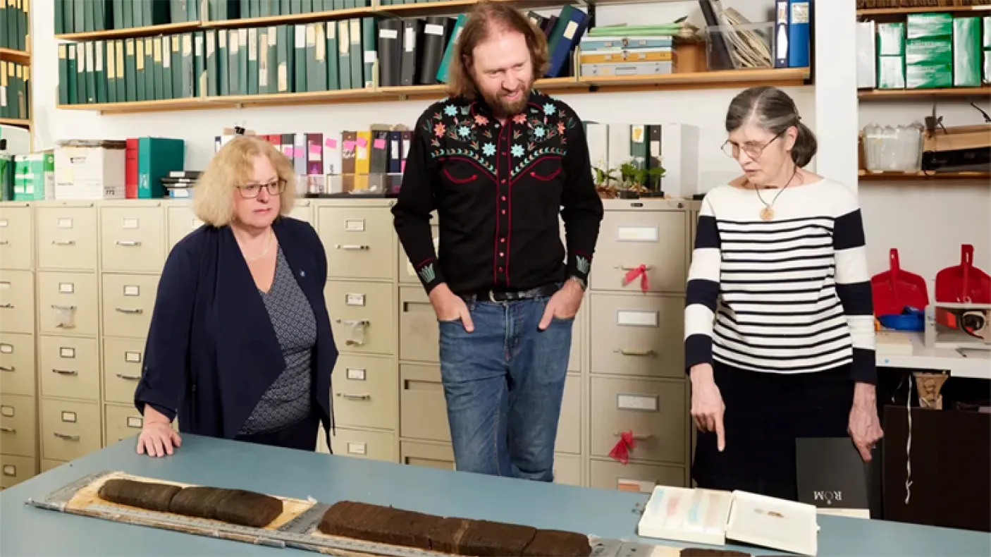 Three people standing behind a table with artifacts and documents in a room filled with shelves and filing cabinets.