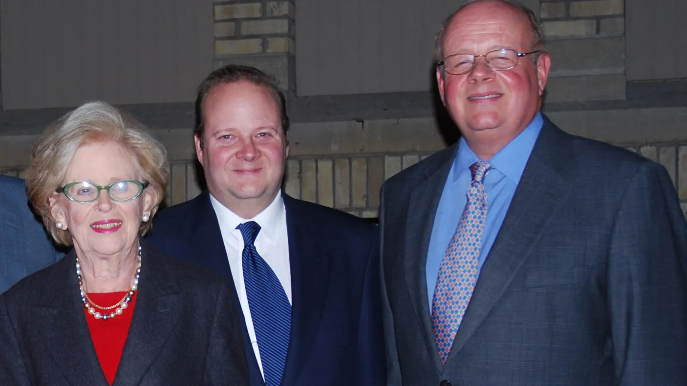 Three people posing closely together, all wearing formal attire against a neutral indoor background.