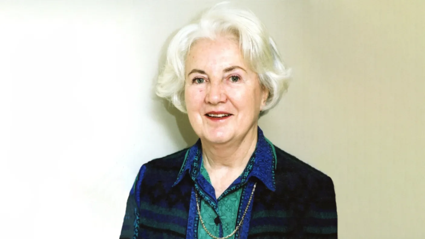 Portrait of an older woman with white hair, wearing a dark vest and a blue patterned blouse against a white background.