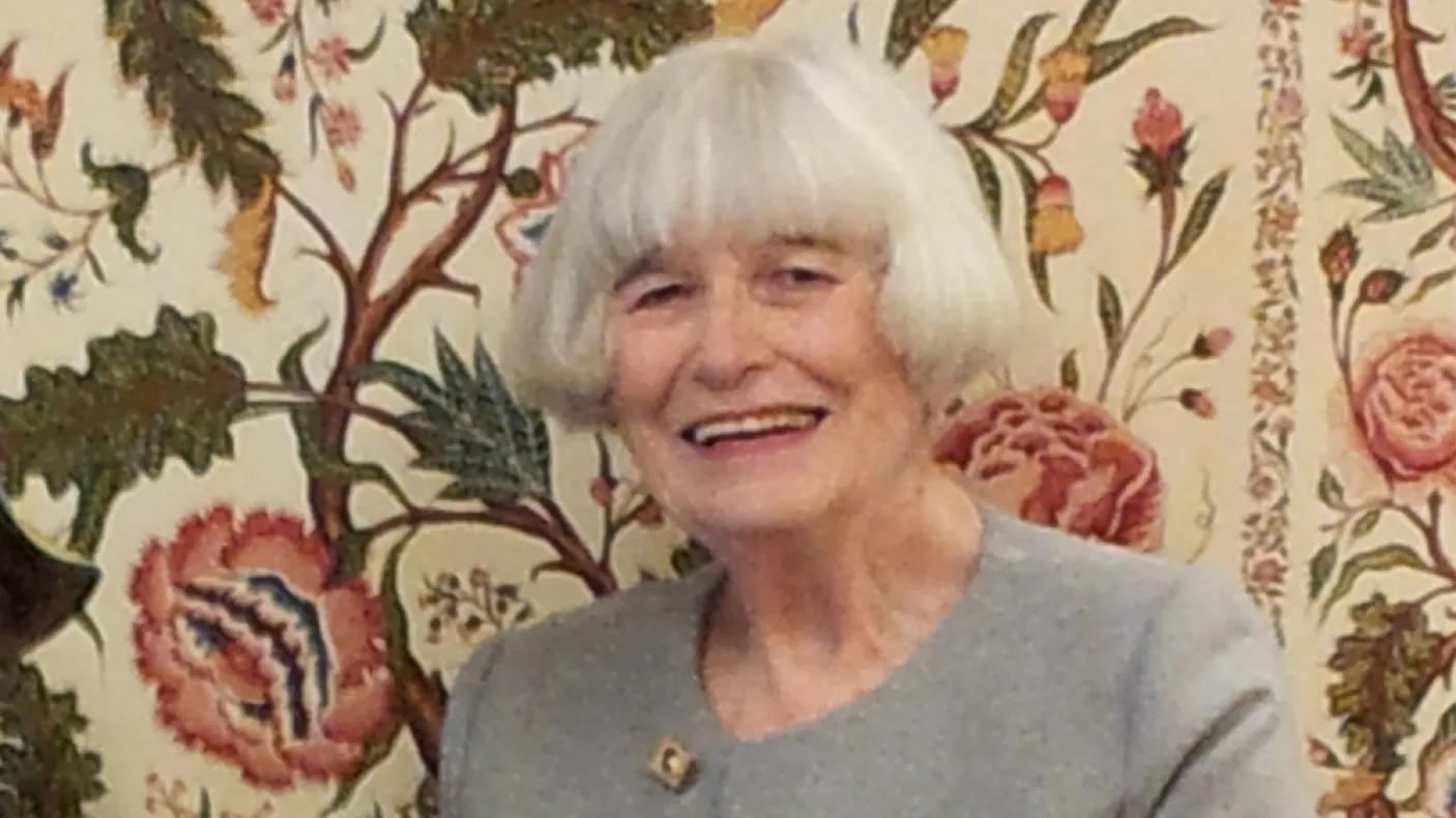 Elderly woman with white hair smiling in front of a floral patterned background.