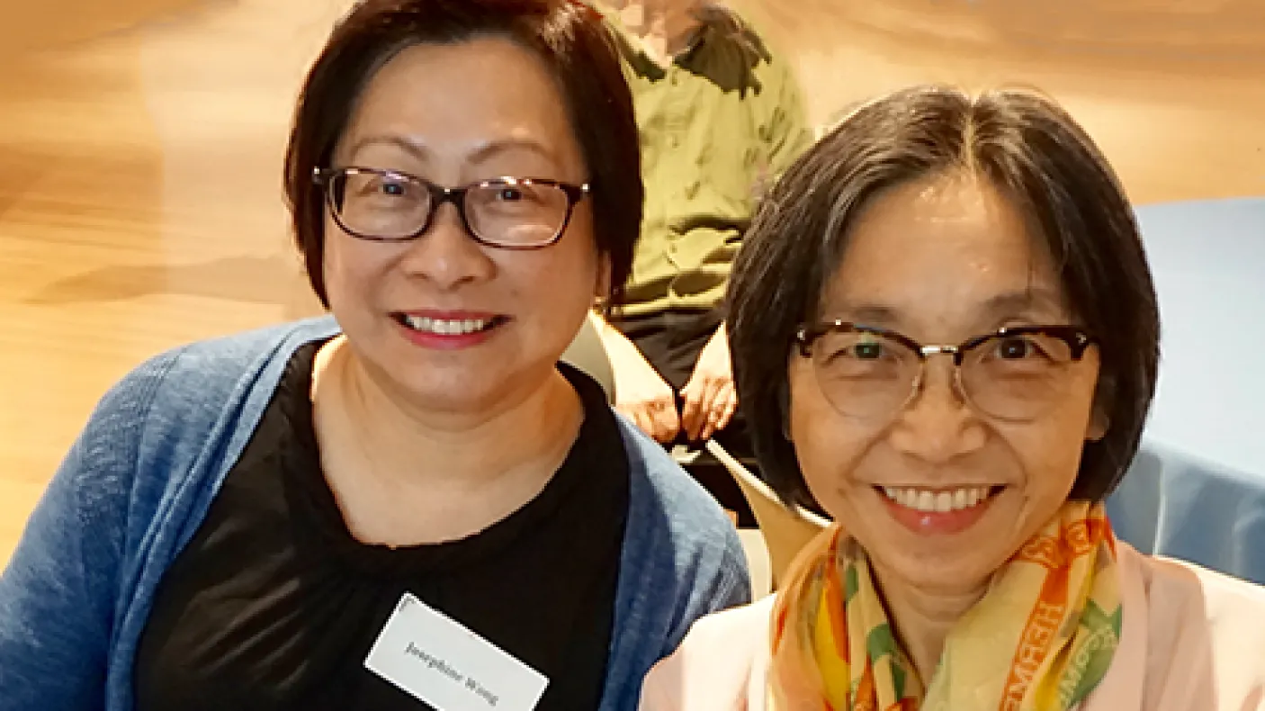 Two smiling women wearing glasses, seated indoors with one wearing a name tag.