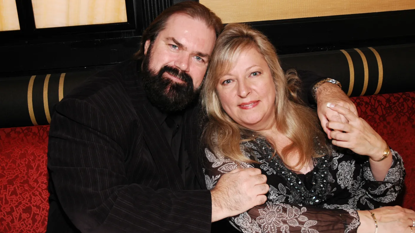 A man with a beard and a woman with blonde hair sit together on a striped couch, both looking at the camera.