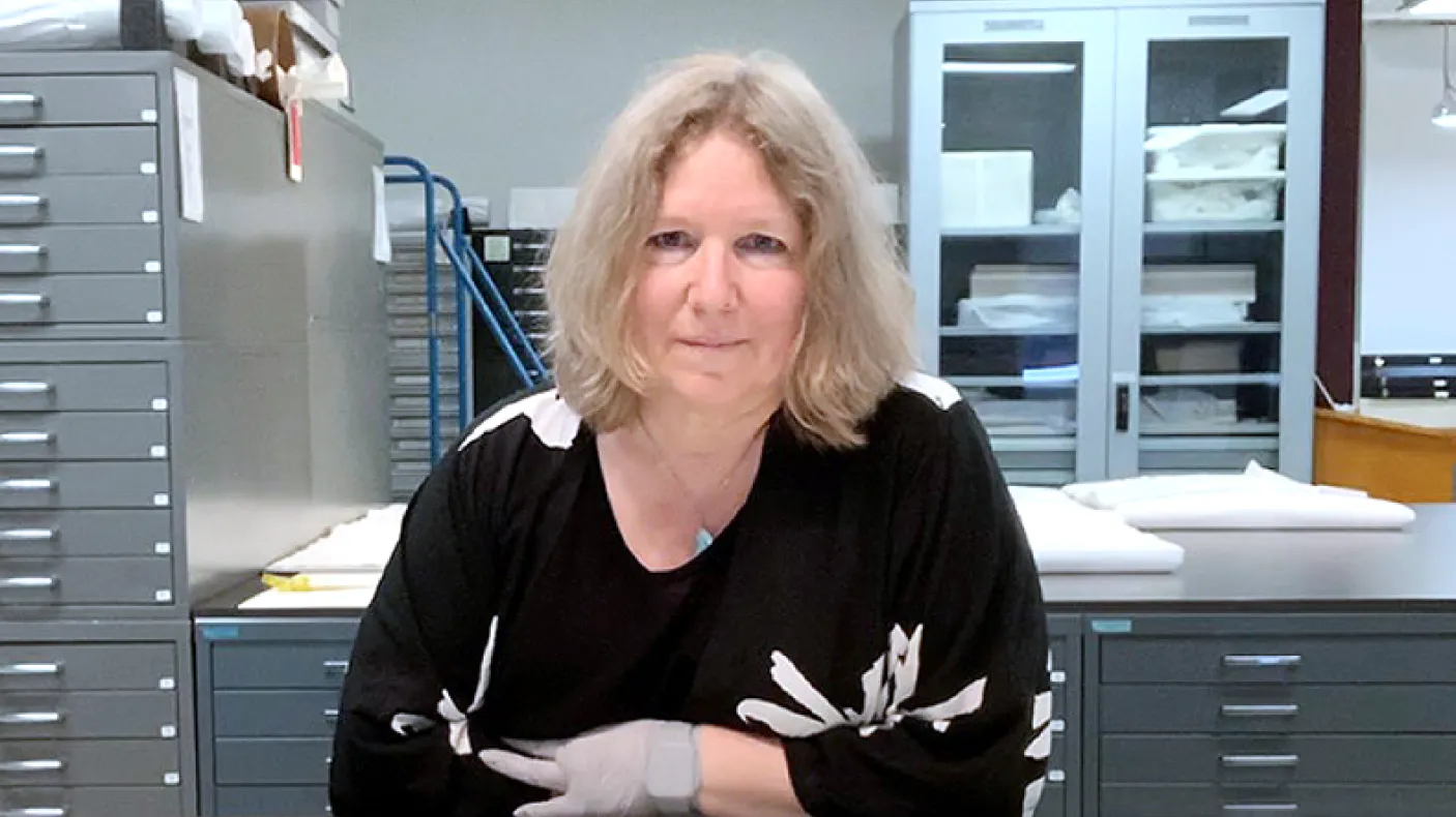A woman with shoulder-length blonde hair wearing a black sweater with white skeleton designs sits at a desk in a laboratory or office setting.