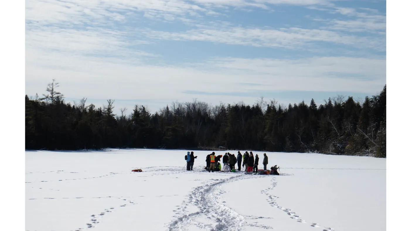 Groupe de personnes rassemblées sur un lac gelé et enneigé, entouré d'arbres, sous un ciel partiellement nuageux.