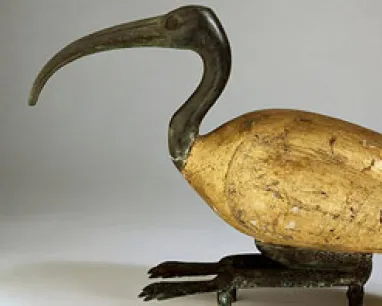 Wooden bird sculpture with a long curved beak and worn yellow and black paint.