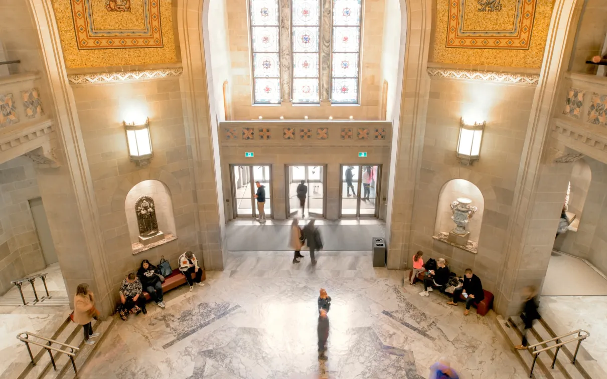 Spacious museum entrance hall with high arched ceiling, detailed wall carvings, and symmetrical seating areas along the sides.