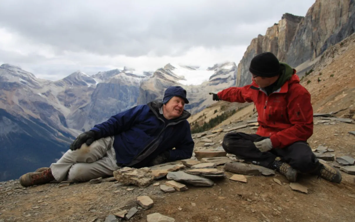 Two people sit and gesture on a rocky mountain trail with towering cliffs and distant peaks in the background.