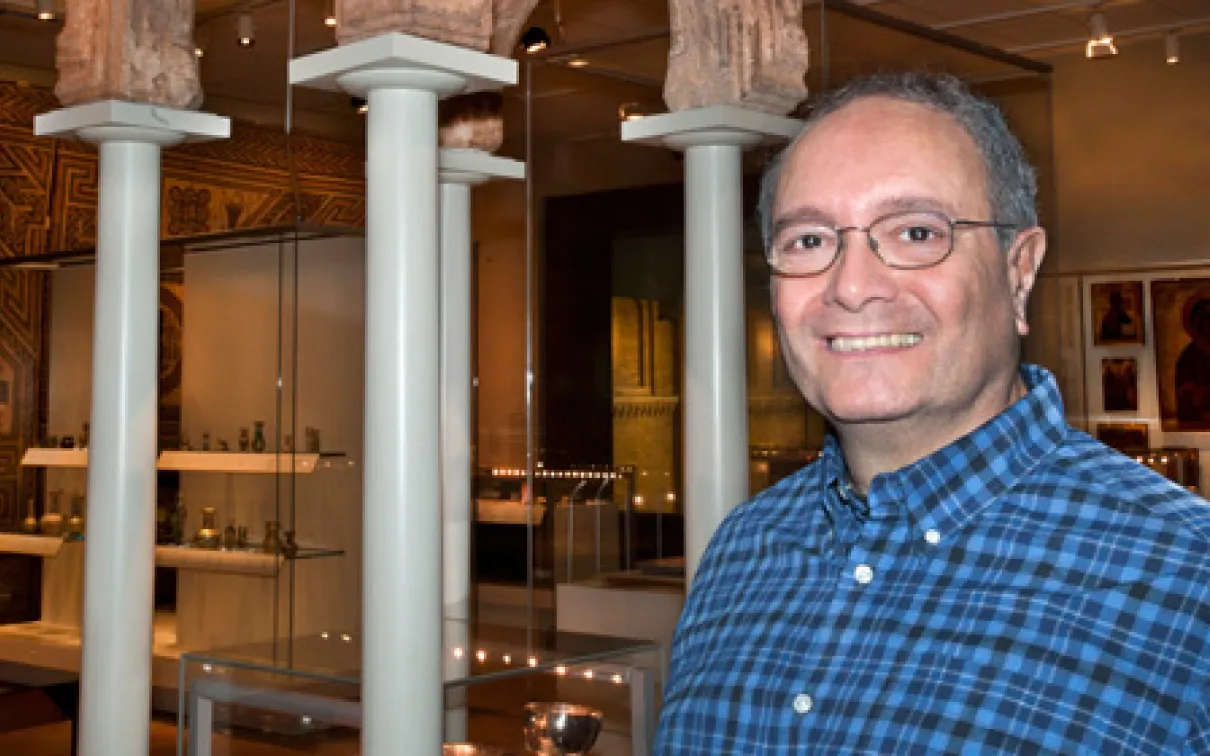 A man wearing glasses and a blue checked shirt stands in a museum gallery with white columns and display cases behind him.