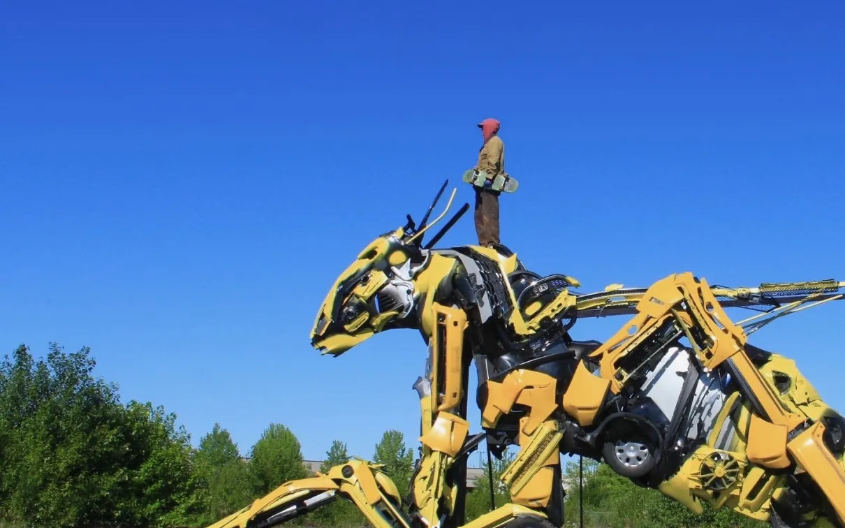 Grande sculpture de dinosaure mécanique jaune fabriquée à partir de pièces industrielles sur une plate-forme en bois, en plein air, sous un ciel bleu clair.