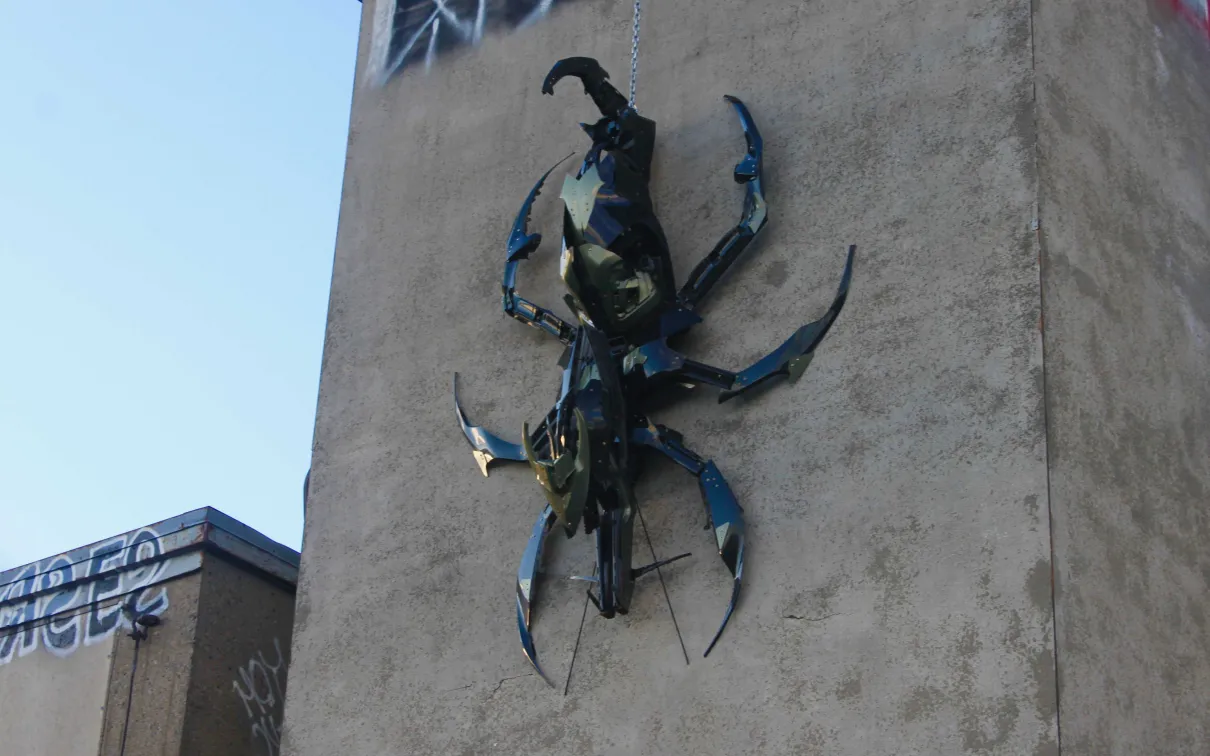 Grande sculpture de scarabée en métal bleu et noir montée sur le mur d'un bâtiment en béton beige.