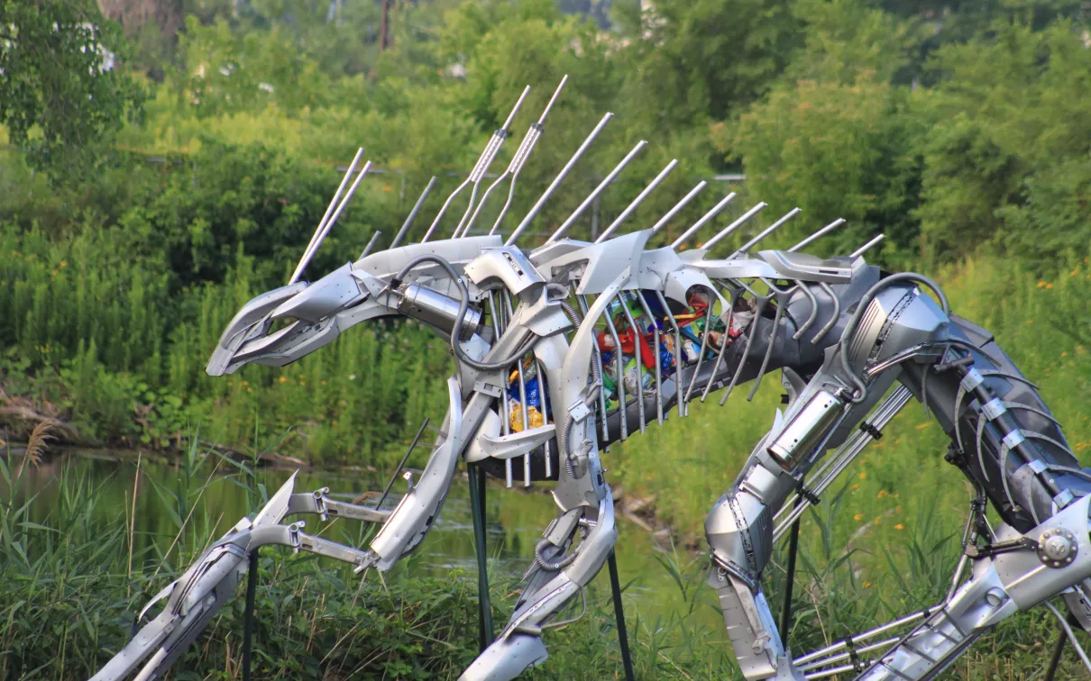 Grande sculpture métallique d'un insecte aux multiples pattes et antennes, placée en extérieur avec de la verdure en arrière-plan.