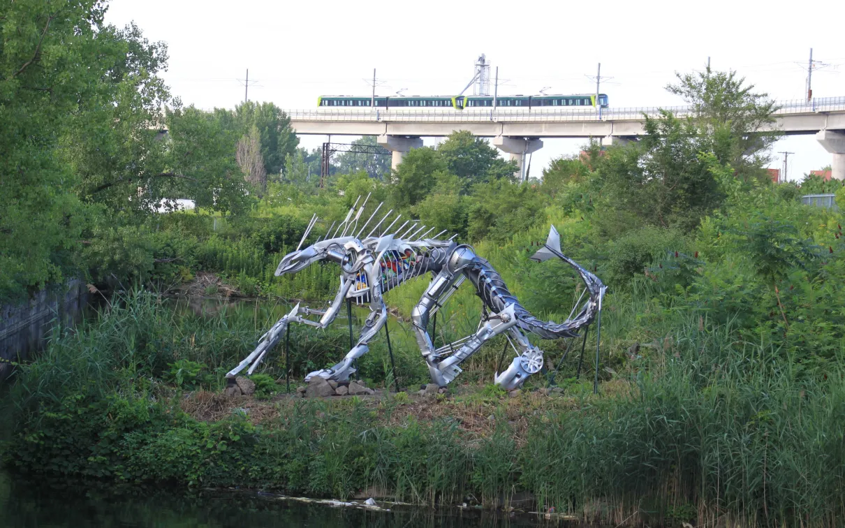 Sculpture en acier représentant un chat à dents de sabre accroupi, entouré de verdure, près d'un plan d'eau, avec un train qui passe à l'arrière-plan.