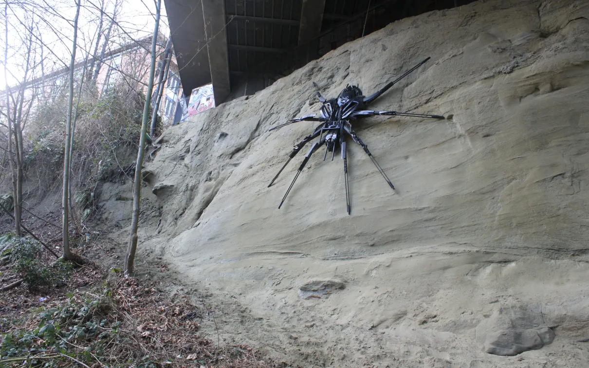 Grande sculpture d'araignée noire montée sur un rocher sablonneux en plein air parmi des arbres dénudés.