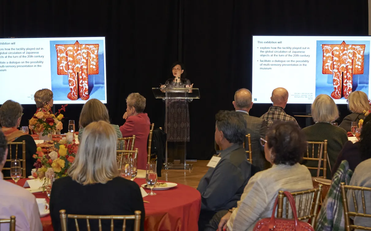 A speaker at a podium presents to an audience seated at round tables with two large screens displaying an artifact and text.