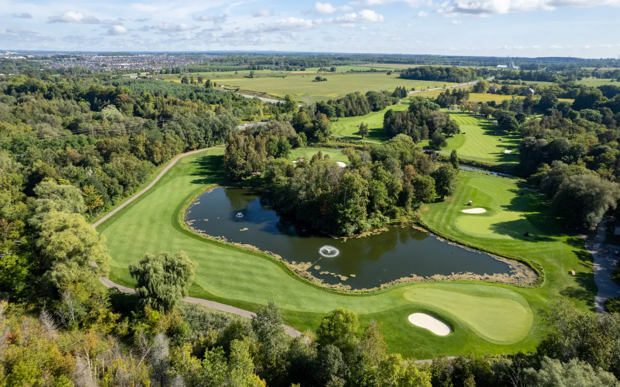Aerial view of a golf course with a water hazard, sand traps, and surrounding trees under a blue sky.