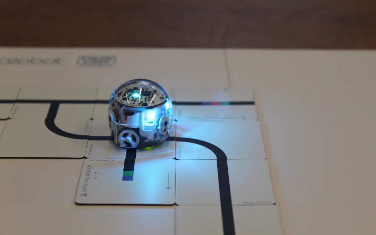 A small round robot with blinking lights follows a dark path along a paper
