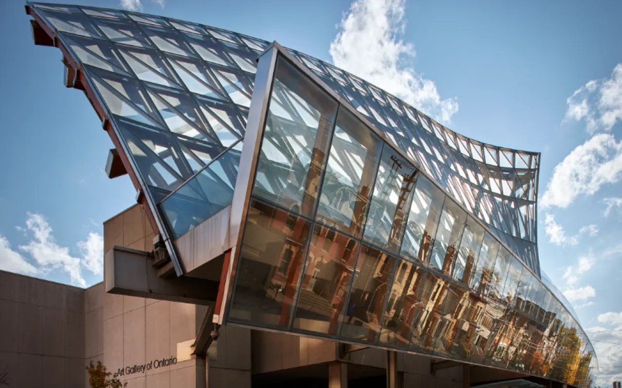 Exterior of the Art Gallery of Ontario featuring a striking curved glass façade with steel framing, reflecting nearby buildings under a blue sky.