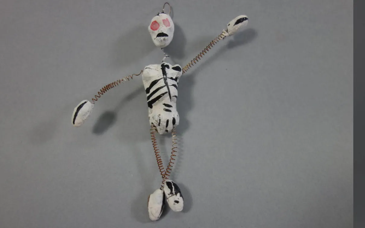 Petite figurine artisanale au corps blanc peint de marques noires, à la bouche rouge et aux yeux orange, posée sur un fond gris.