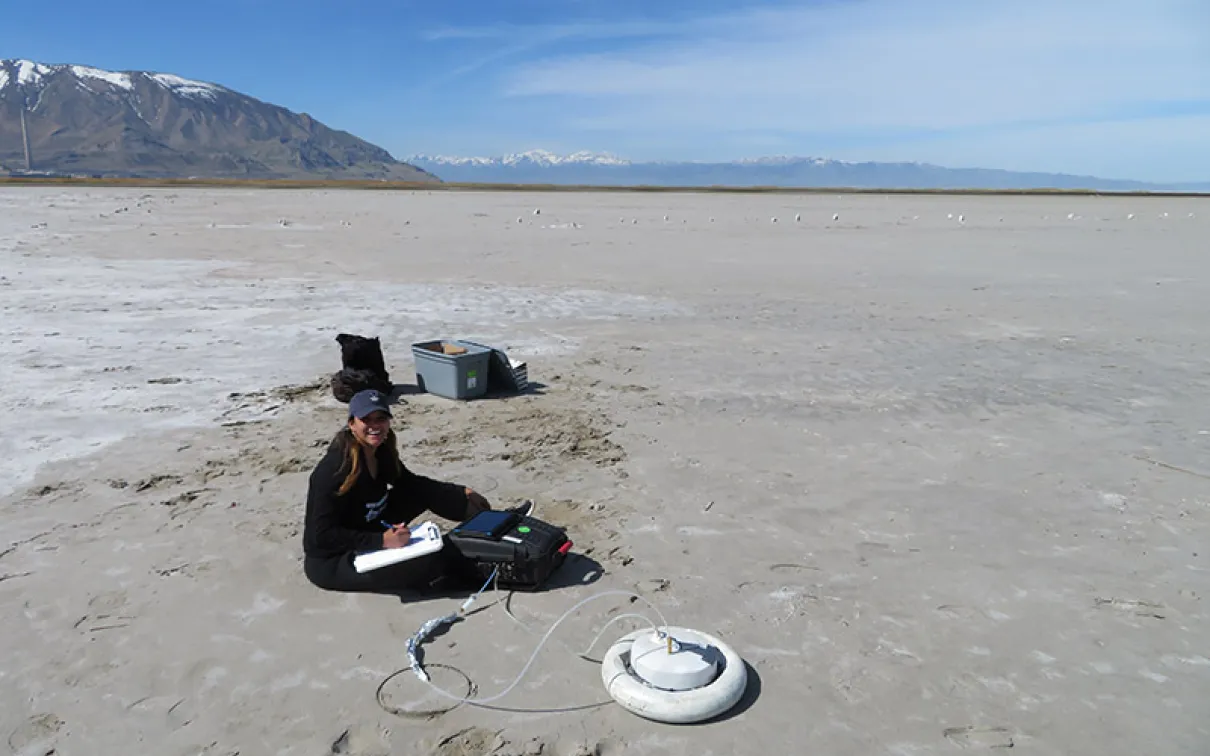Researcher sitting on a dry lakebed next to scientific equipment including a circular white sensor device.