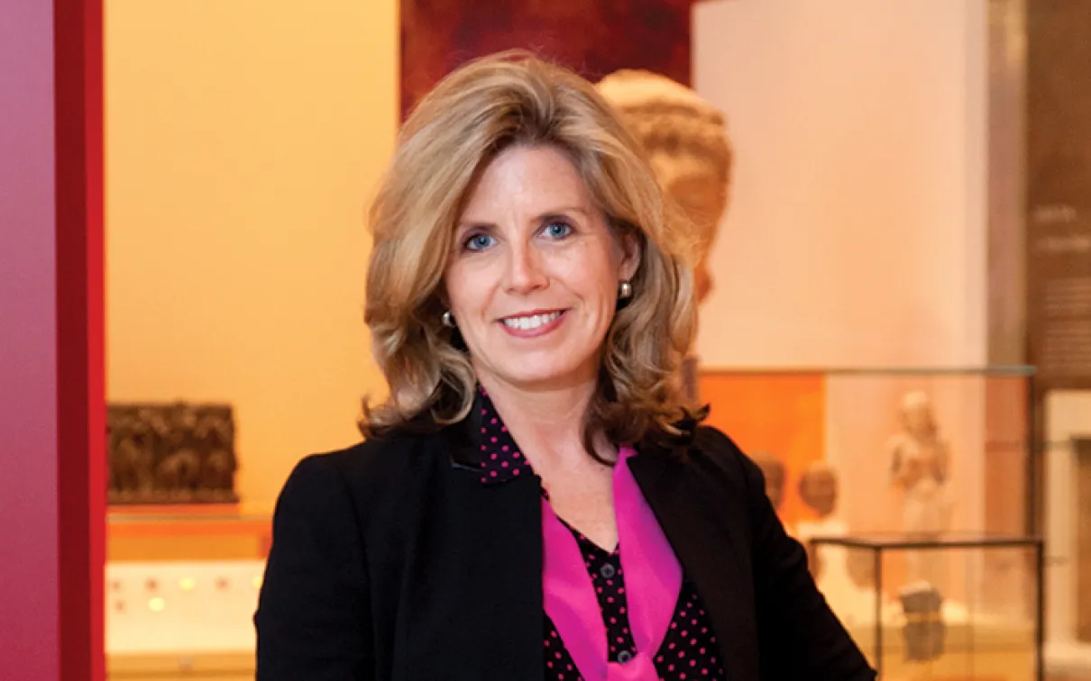 Woman with shoulder-length blonde hair wearing a black blazer over a pink blouse, standing indoors with blurred background.