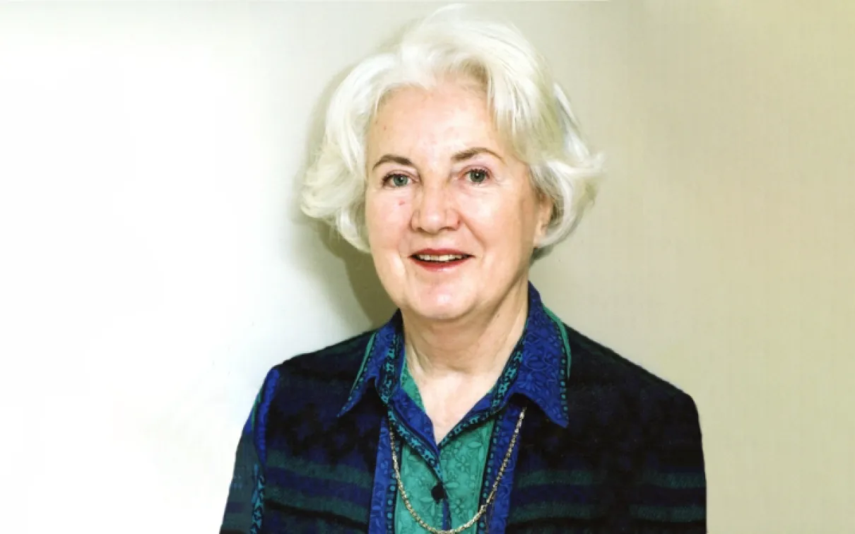 Portrait of an older woman with white hair, wearing a dark vest and a blue patterned blouse against a white background.