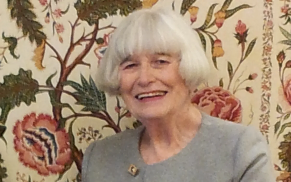 Elderly woman with white hair smiling in front of a floral patterned background.