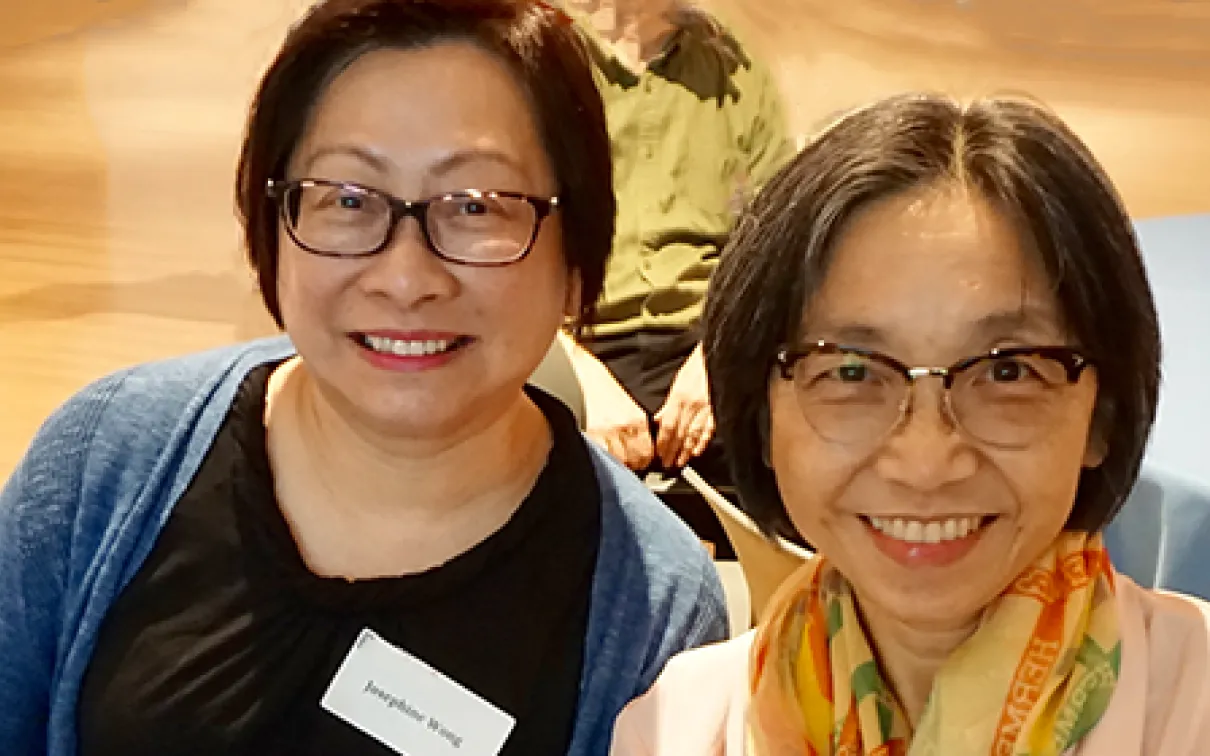 Two smiling women wearing glasses, seated indoors with one wearing a name tag.