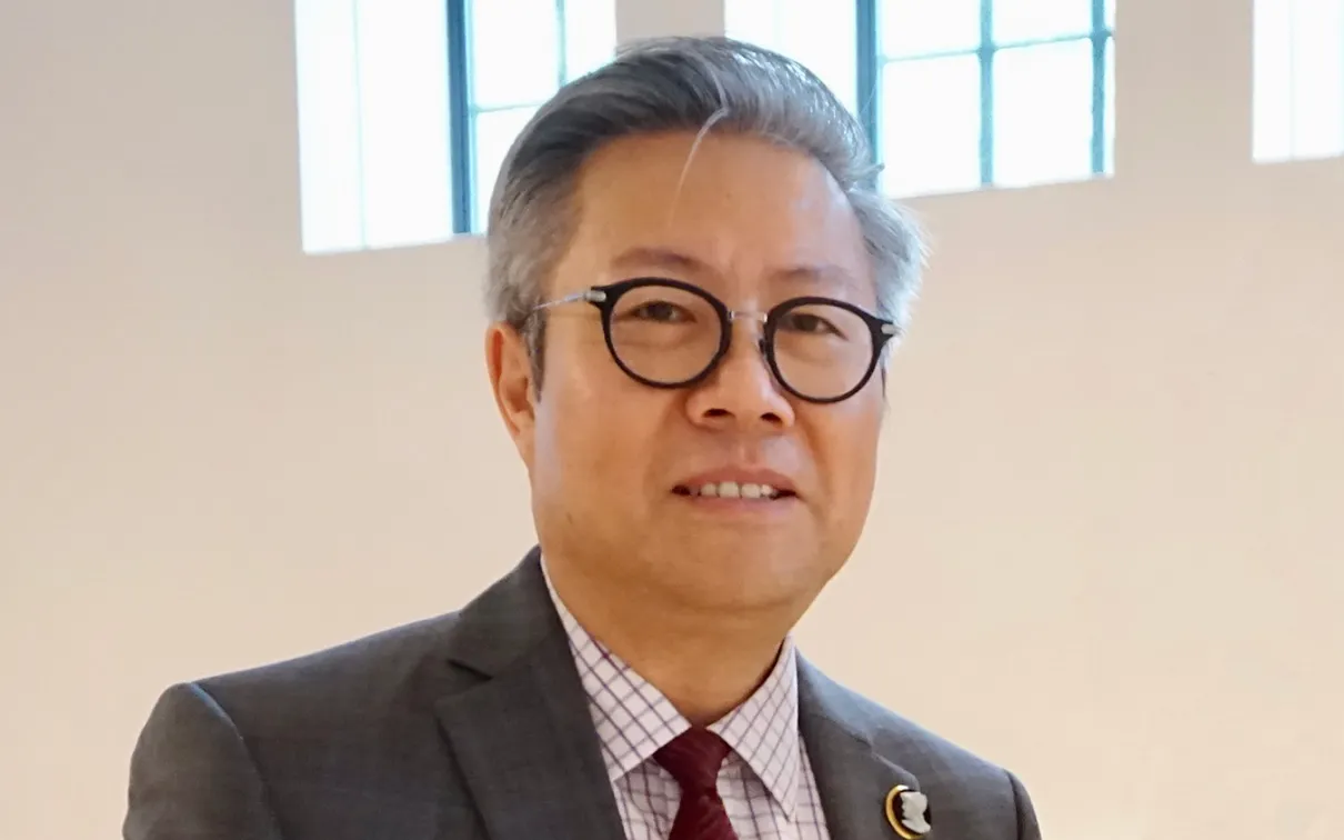 Man with glasses wearing a dark suit and red patterned tie, standing indoors against a beige wall.