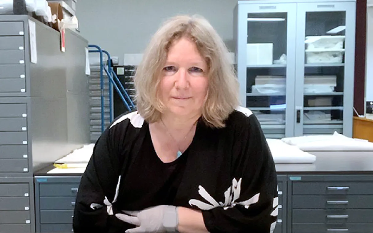 A woman with shoulder-length blonde hair wearing a black sweater with white skeleton designs sits at a desk in a laboratory or office setting.