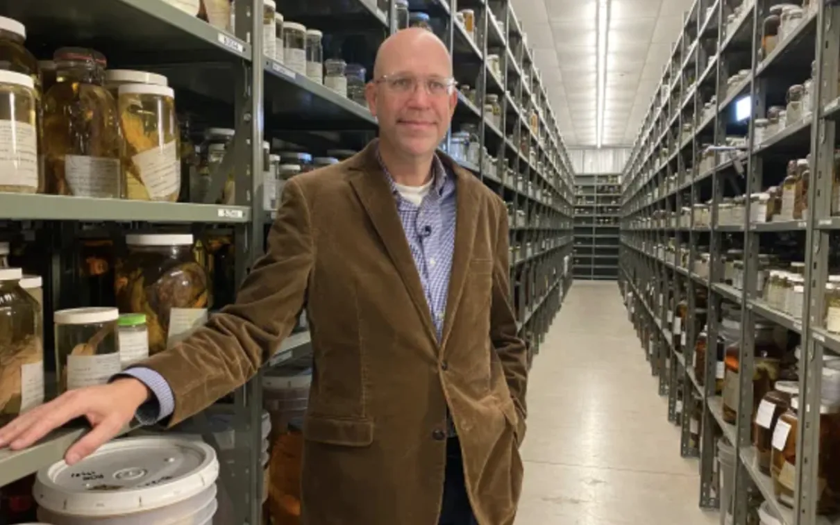 A man wearing glasses, a brown jacket, and a blue shirt stands in a narrow aisle lined with shelves stocked with jars and containers.