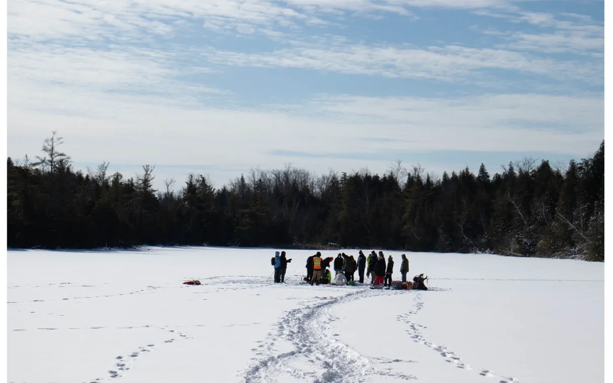 Groupe de personnes rassemblées sur un lac gelé et enneigé, entouré d'arbres, sous un ciel partiellement nuageux.