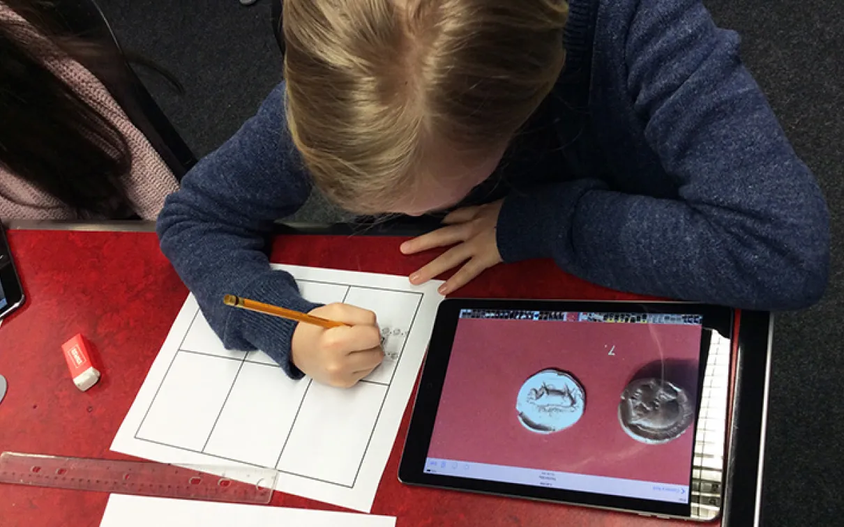 A student draws on a piece of paper with a coin image open on her iPad
