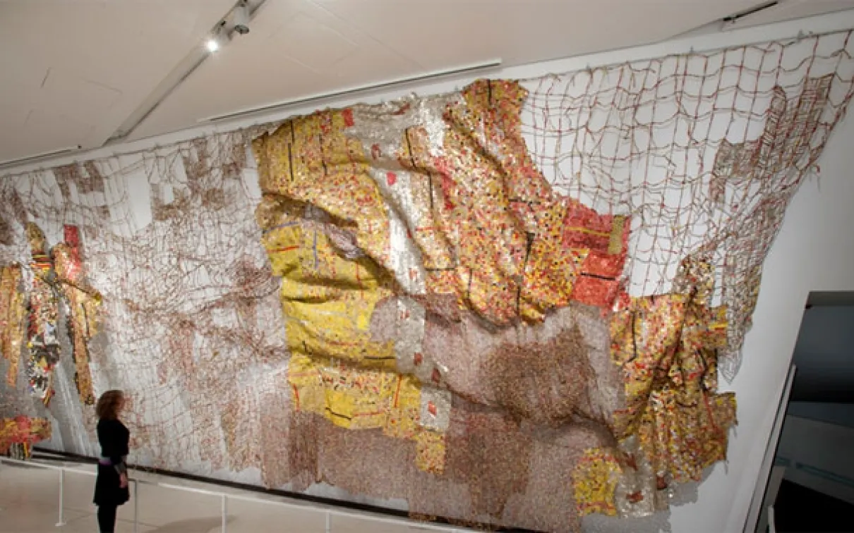 Large wall-mounted map artwork made from netting, layered fabrics, and various textures in brown, yellow, orange, and red tones.