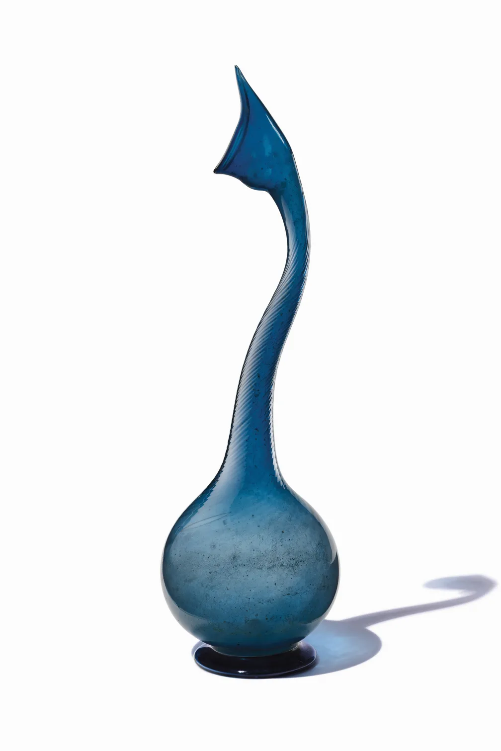 Tall, slender blue glass vase with a curved neck and bulbous base casting a shadow.