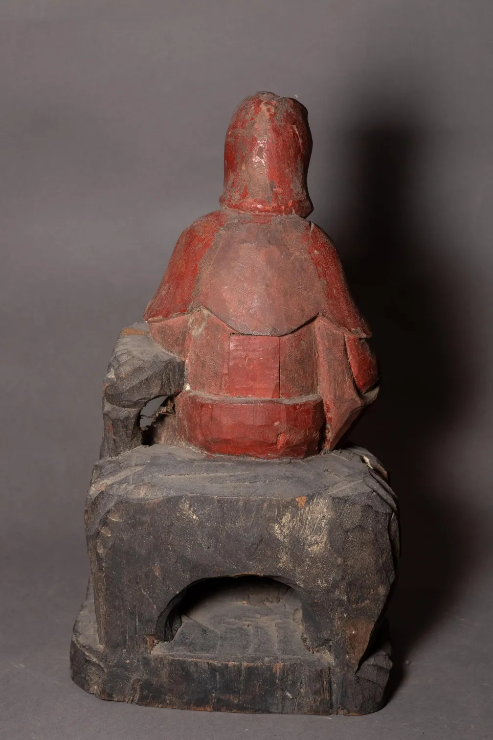 Wooden sculpture painted red and black depicting a seated figure with a rounded back and covered head.