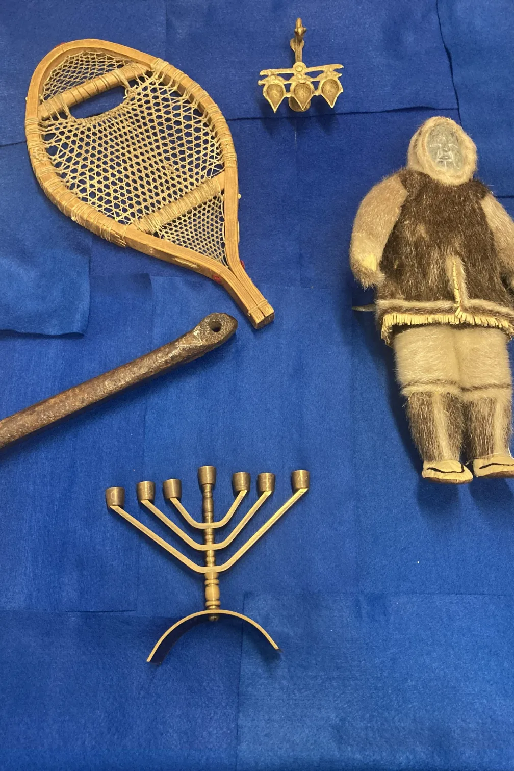 Sélection d'objets sur fond bleu.  Les objets comprennent une poupée inuit en pierre et en peau de phoque, une Hannukiah, un patin médiéval en os, une raquette et une lampe diya.