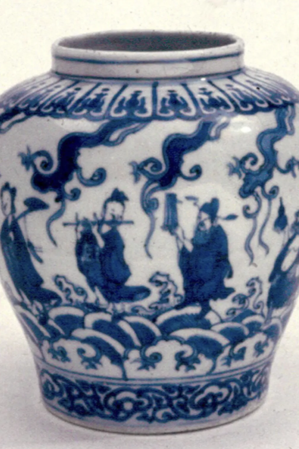 Blue-and-white ceramic jar with a wide mouth and rounded body. The surface is decorated with cobalt-blue painted scenes of several robed figures standing among stylized waves and swirling cloud-like shapes. Decorative bands of repeating patterns encircle the rim and base. The jar is photographed against a plain, light background, highlighting the contrast between the white glaze and the deep blue designs.