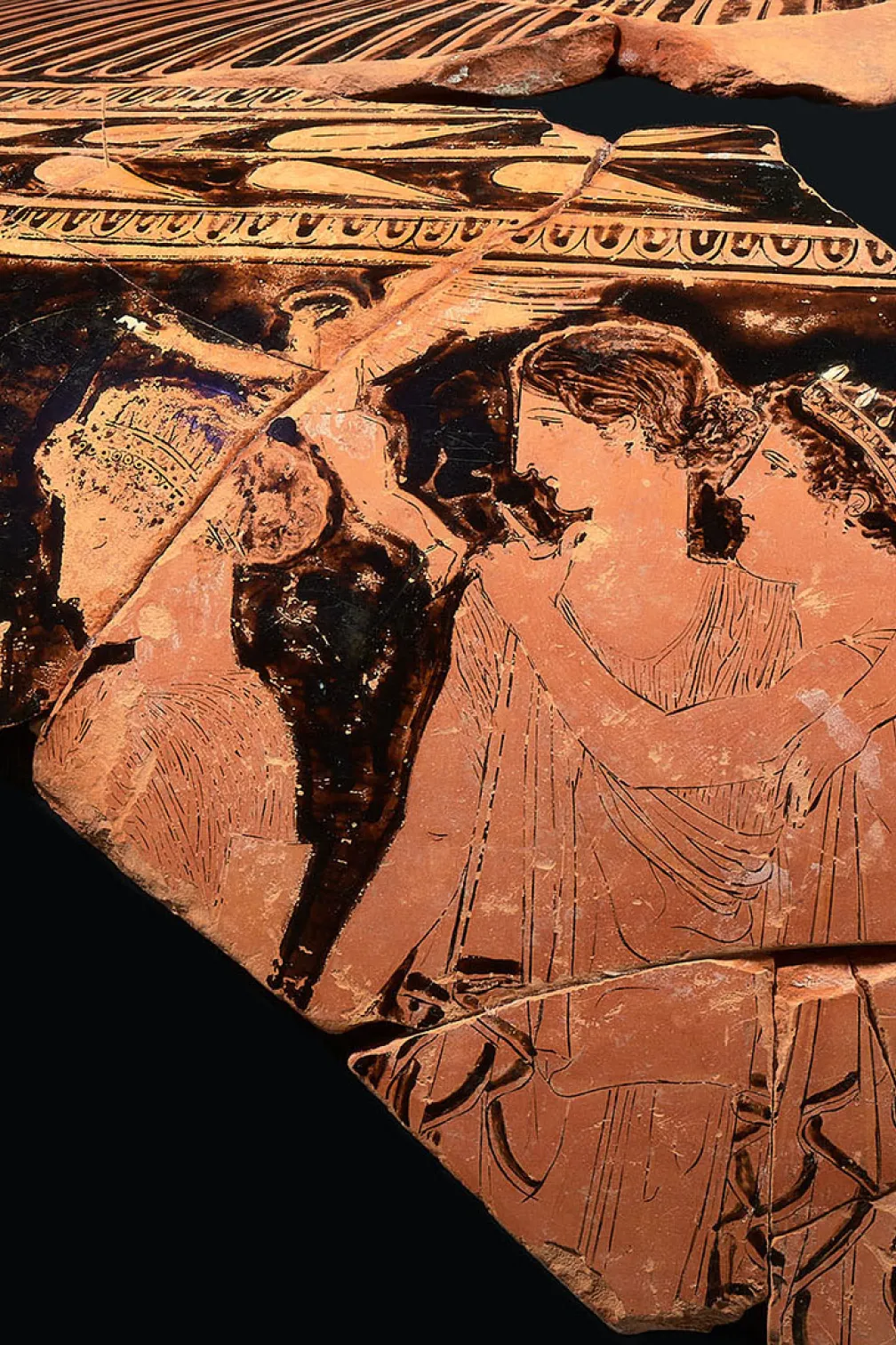 Fragment de poterie grecque antique en terre cuite représentant deux personnages de profil avec des motifs décoratifs au-dessus.