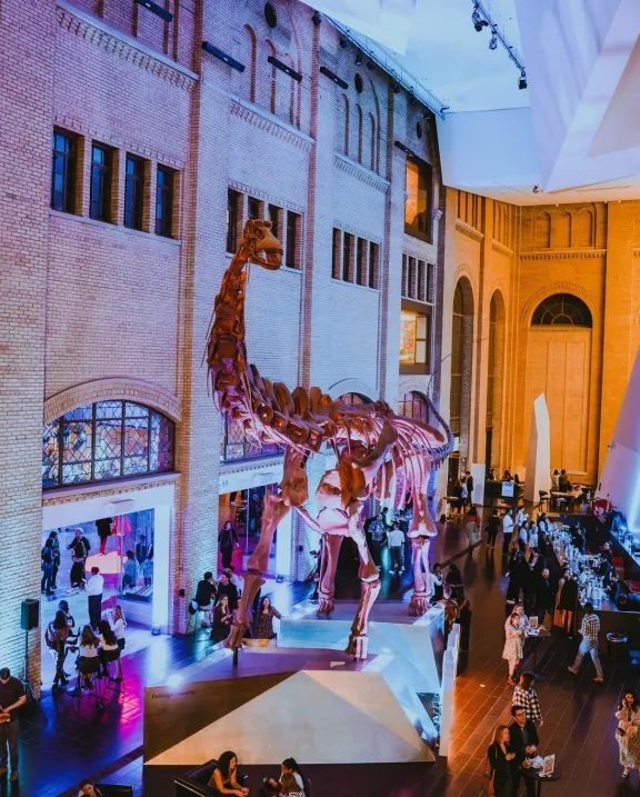 Interior view of a museum hall with visitors, featuring dinosaur skeleton displays and illuminated architectural columns.