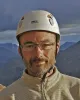 Man wearing glasses and a white Petzl helmet with a cloudy sky and mountains in the background.