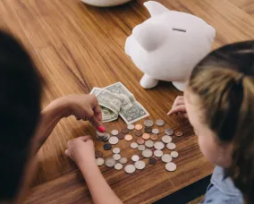 Two children placing coins into a white piggy bank on a wooden table.