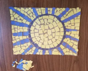 Une mosaïque de soleil presque complète est posée sur une table. Un anneau bleu foncé et des rayons bleu roi représentent le soleil. Le centre du soleil est rempli de jaune. Les carrés bleus et jaunes non utilisés sont empilés à côté de la mosaïque.