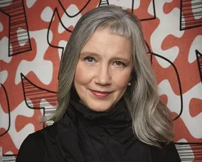 Woman with long gray hair wearing a black turtleneck stands in front of a red and white abstract patterned wall.