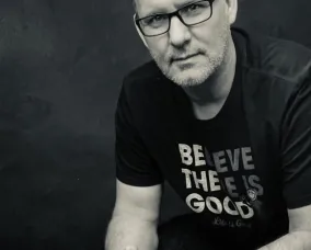 Homme aux cheveux gris courts et aux lunettes portant une chemise noire avec le texte blanc "BELIEVE THERE IS GOOD".