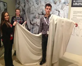 The young women hold the fabric on the young man’s white side. The fabric passes beneath the young man’s arm.